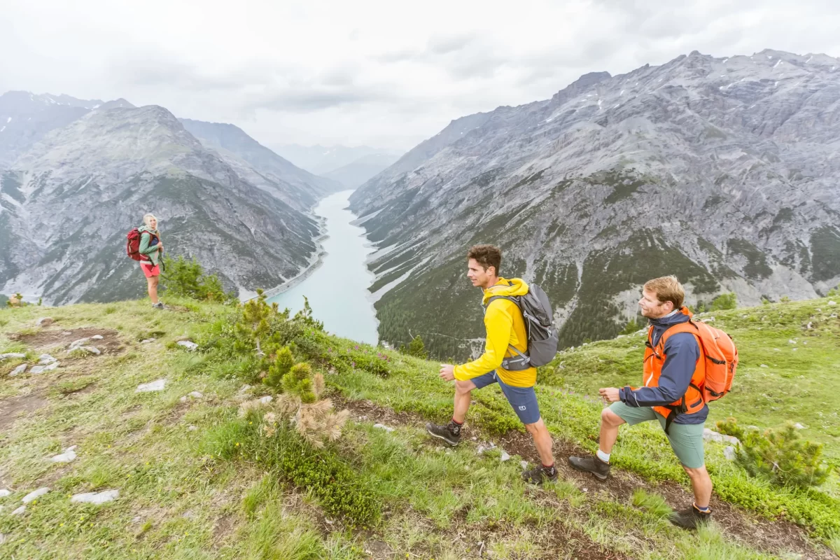 Tre escursionisti camminano su un sentiero panoramico di montagna, con un lago e montagne sullo sfondo.