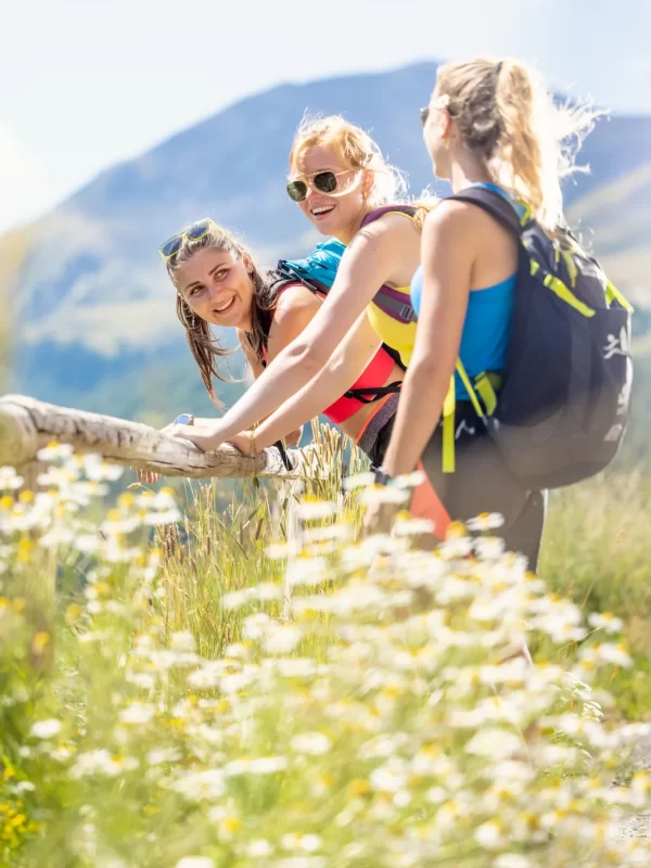 Tre donne sorridenti fanno escursioni in montagna circondate da fiori selvatici.