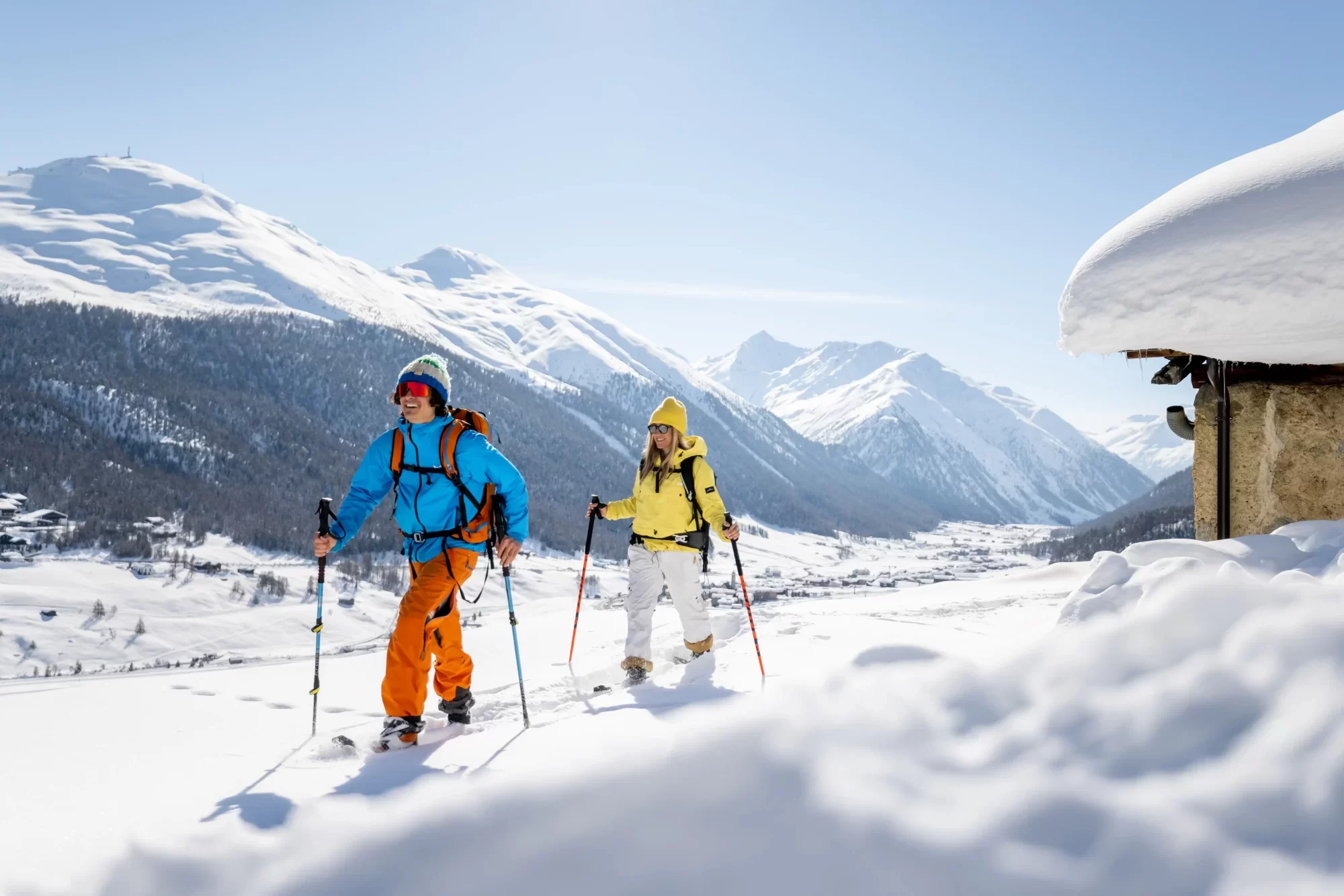 Due persone fanno sci alpinismo su un pendio innevato con montagne sullo sfondo.
