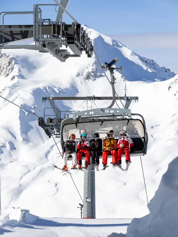 Sciatori su una seggiovia con montagne innevate sullo sfondo.