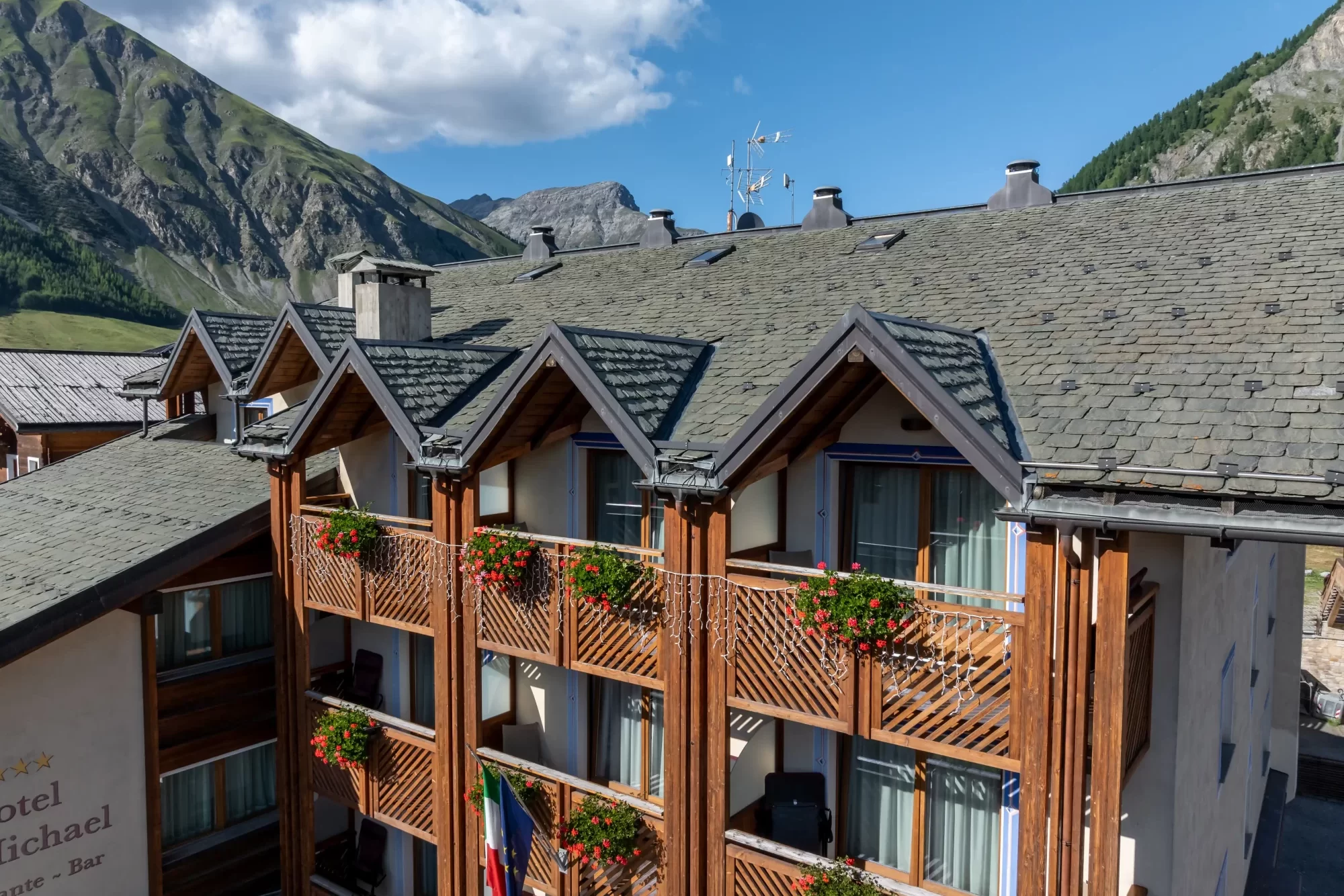 Facciata di un hotel di montagna con balconi in legno e fiori, circondato da montagne.