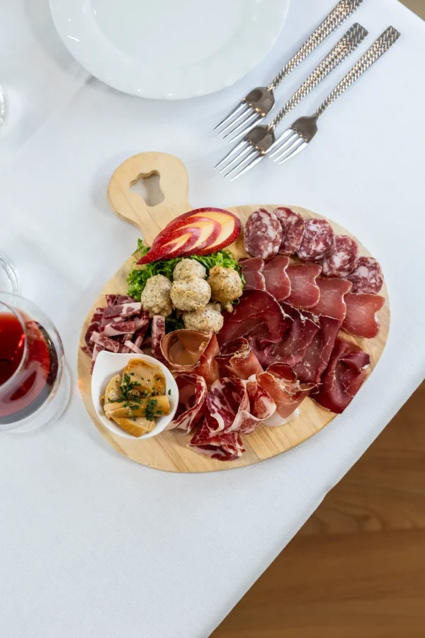 Tagliere di salumi variato con contorno di funghi e un bicchiere di vino rosso su tavola apparecchiata.