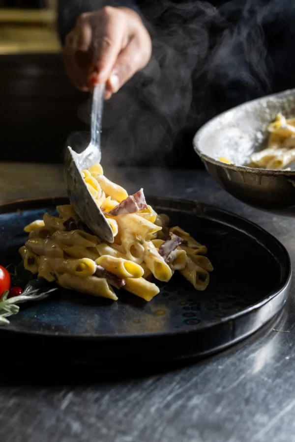 Una persona serve penne cremose con pancetta su un piatto nero.