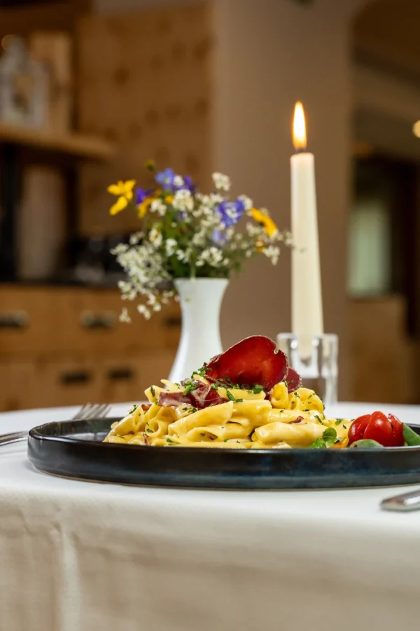 Piatto di pasta cremosa con prosciutto su tavola apparecchiata elegantemente con candela accesa e vaso di fiori.