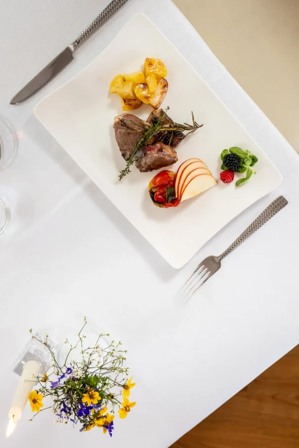 Piatto gourmet con carne, patate, frutta e verdure su una tovaglia bianca.