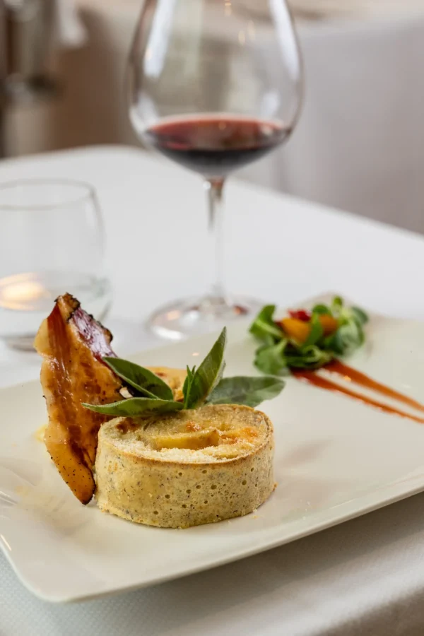 Piatto gourmet con tortino salato, pancetta croccante e insalata, accompagnato da un bicchiere di vino rosso.