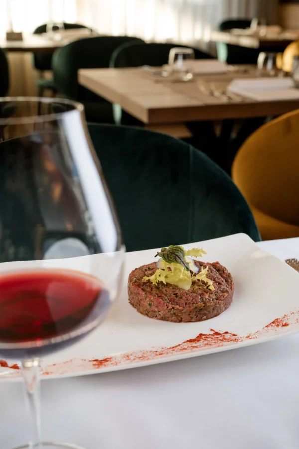 Tartare di carne con decorazione di foglie su un piatto bianco accanto a un calice di vino rosso in un ristorante elegante.