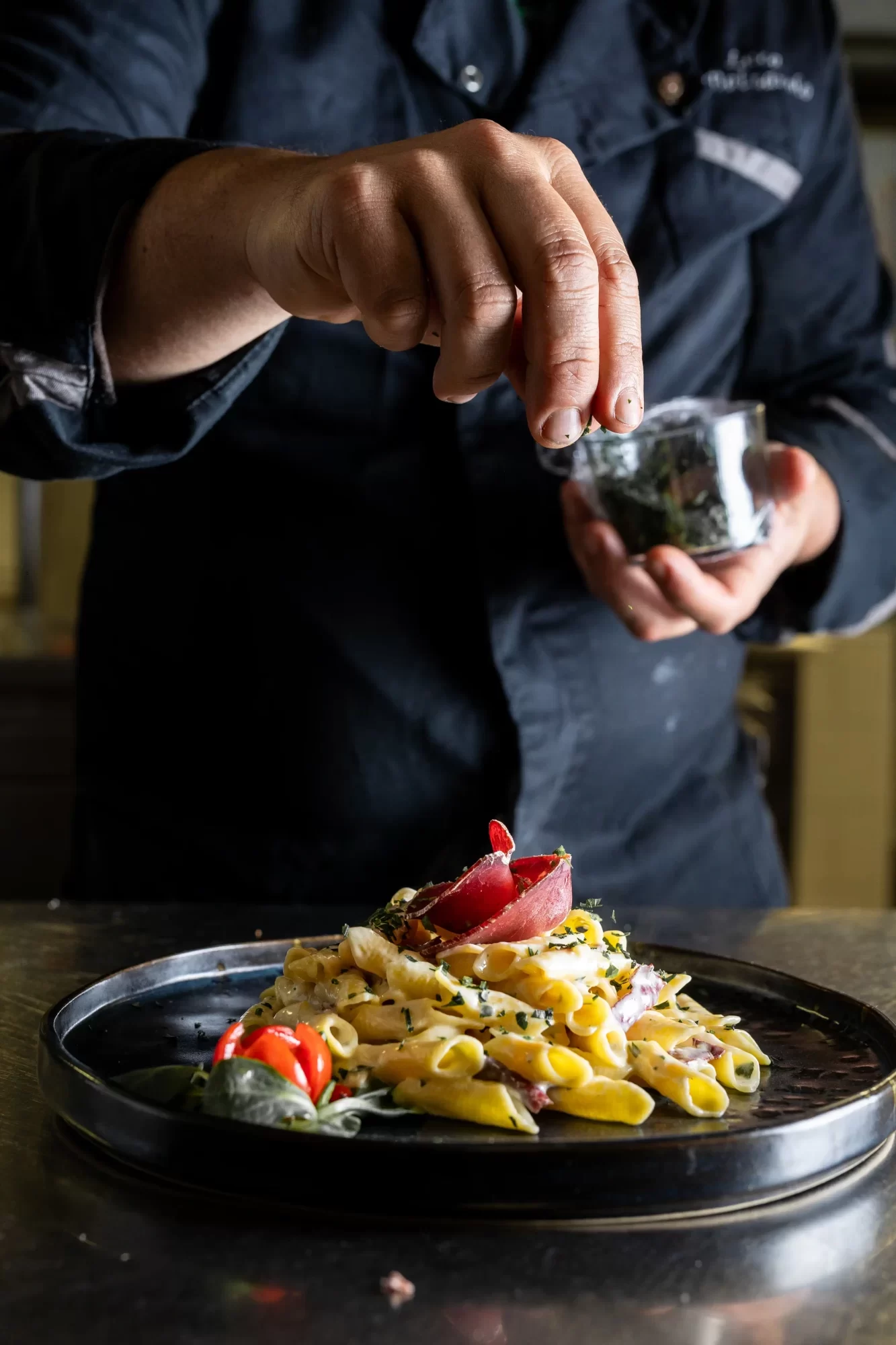 Cuoco aggiunge erbe aromatiche su un piatto di pasta con carne e pomodorini.